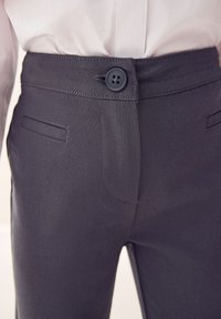 Sivé nohavice s hladkou textúrou, vybavené zapínaním na gombík, pútkami na opasok a bočnými vreckami. Strih na mieru, detailné prešívanie.