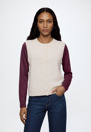 Donna con capelli lunghi e scuri che indossa un gilet beige abbottonato sopra una camicia a maniche lunghe color granata e jeans blu, in piedi con una mano in tasca.