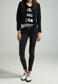 Chaqueta de mezclilla negra sobre una camiseta gráfica, combinada con jeans skinny negros y zapatos blancos de cordones. Textura suave, diseño ajustado.