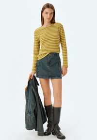 Gestreifte Senf-Langarmbluse, blauer Jeansrock, schwarze kniehohe Stiefel, hält eine Jeansjacke mit braunen Akzenten.