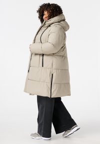 Manteau matelassé beige avec une capuche, présentant des motifs matelassés et des accents de fermeture éclair noirs. Associé à un pantalon noir à jambes larges et des baskets grises.