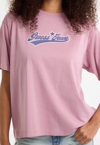 Nais, kes kannab heledat roosa Guess Jeans t-särki sinise logo tekstiga, kombinatsioonis heledate siniste teksadega, käsi puusal.