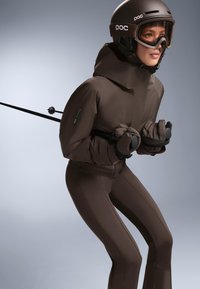 Lyžiarsky overal v tmavohnedej farbe so priliehavým strihom, obsahujúci vysoký golier, zipsové vrecká a rukavice. Doplnky zahŕňajú prilbu a lyžiarske okuliare.