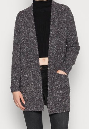 Persoon draagt een donkergrijs, openvallend vest met structuur over een zwart, kort coltrui-trui en een zwarte broek, met één hand in de zak.