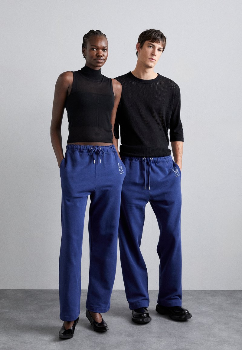 Lässiges Outfit bestehend aus einem schwarzen ärmellosen Oberteil, blauen Sweatpants mit Tunnelzug und Seitentaschen sowie kontrastierenden schwarzen Schuhen.