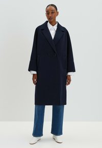 Marineblauer Doppelreiher-Mantel mit Reverskragen, Seitentaschen und breiten Ärmeln; kombiniert mit einem weißen Hemd und blauen weit geschnittenen Jeans.