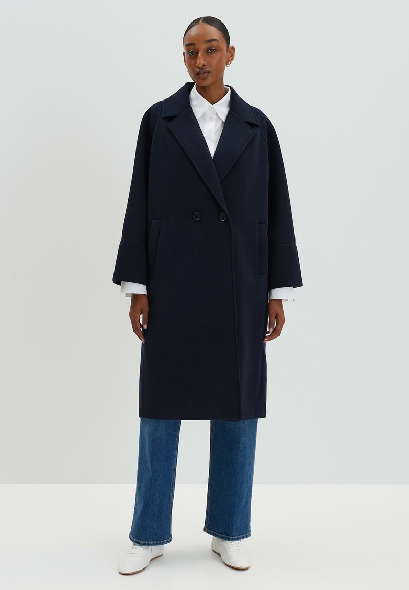 Marineblauer Doppelreiher-Mantel mit Reverskragen, Seitentaschen und breiten Ärmeln; kombiniert mit einem weißen Hemd und blauen weit geschnittenen Jeans.