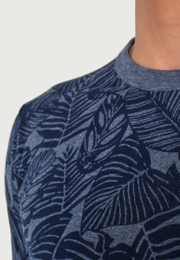 Gros plan sur une personne portant un pull à col rond bleu avec un motif de feuilles tropicales bleu foncé sur le tissu.