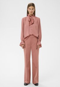 Rynket pink bluse med sløjfedetalje, lange ærmer og udsvingede manchetter; parret med matchende lyserøde bukser med vidde. Sorte sko.