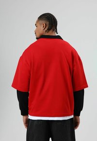 Rode oversized poloshirt met een zwarte kraag, korte mouwen en een gladde textuur; gedragen over een zwart shirt met lange mouwen en donkere broek.