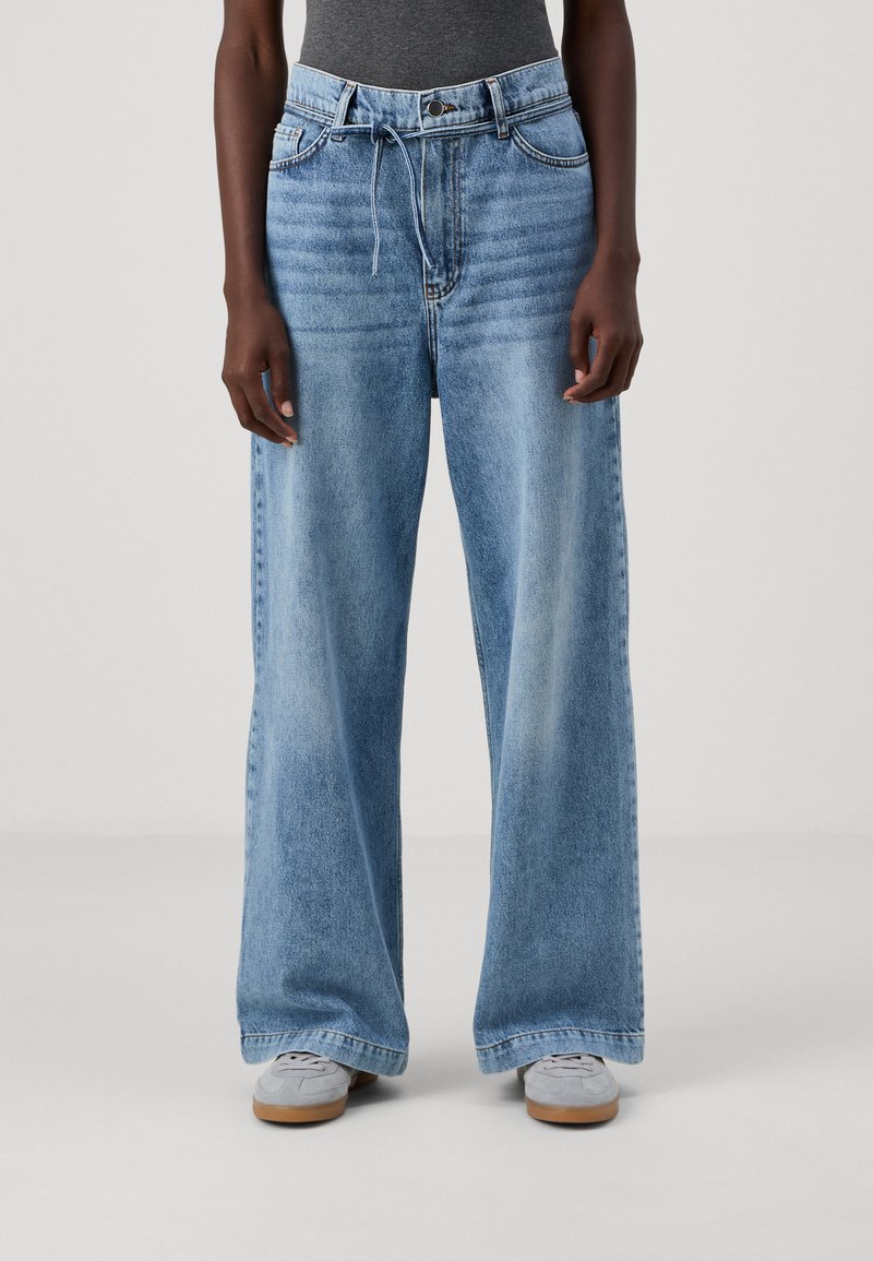 Wide-leg denim jeans in light blue, featuring a drawstring waistband and faded texture. Paired with gray shoes on a plain background.