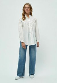 Camicia bianca con design a yoke strutturato, maniche lunghe a sbuffo e colletto, abbinata a jeans blu larghi e sneakers bianche.