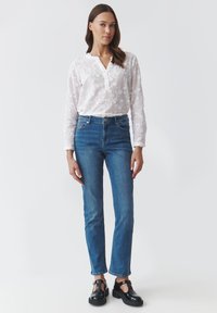 Blusa blanca bordada con un escote abotonado, combinada con jeans de pierna recta azul y zapatos negros de tira en T. Texturas suaves.