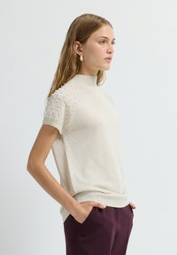 Crème gebreide trui met korte mouwen, hoge halslijn en gemêleerde versieringen op de schouders; gedragen met bordeauxkleurige broek.