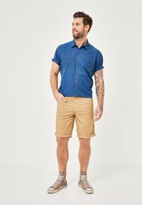 Denim shirt in dark blue with two chest pockets, paired with beige rolled shorts. Gray sneakers complete the outfit. Displayed against a light backdrop.