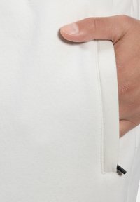 Bílá bavlněná tkanina s viditelnou kapsou. Ruka je vložena do kapsy, což odhaluje švy a malý černý detail.