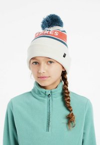 Berretto di maglia bianco con pompon blu, fasce a righe arancio e blu, indossato da un ragazzo con acconciatura a treccia, abbinato a una giacca in pile teal.