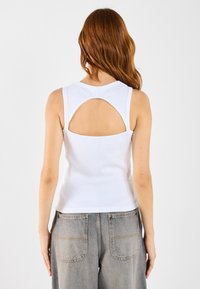 Hvit ermeløs topp med et sirkulært utskjæring bak. Laget av et glatt, elastisk materiale. Kombinert med høyt midje grå jeans.