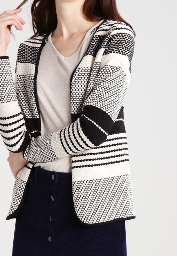 Femme portant un cardigan en maille à motif noir et blanc par-dessus un haut beige clair et une jupe sombre boutonnée, sur un fond uni.
