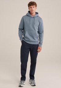 Gaiši zils kapuce ar priekšējo kabatu, regulējamām šņorēm un ribotām piedurknēm, kombinēta ar tumši ziliem biksēm un pelēkiem sporta apaviem.