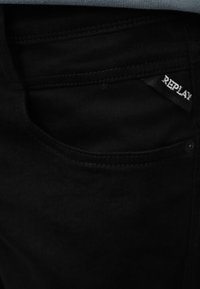 Tissu en denim noir montrant une poche avec des coutures visibles et une petite étiquette noire avec du texte blanc "REPLAY".