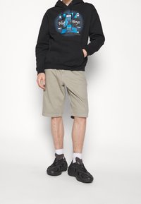 Sudadera negra con estampado gráfico, pantalones cortos beige y zapatos negros. El conjunto presenta un ajuste relajado y un estilo casual adecuado para el clima cálido.
