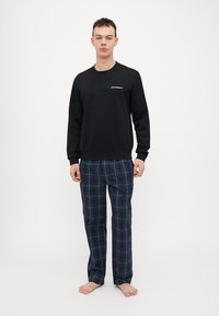 Sweatshirt noir à manches longues avec logo sur la poitrine, associé à un pantalon à carreaux bleu marine. Tissu texturé, coupe décontractée. Pieds nus.