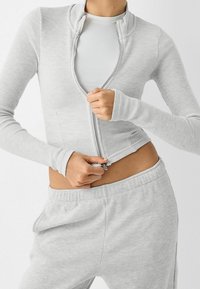 Giacca grigia a zip con coste, aderente con maniche lunghe, abbinata a pantaloni texturizzati coordinati. Il tessuto sembra morbido ed elasticizzato.