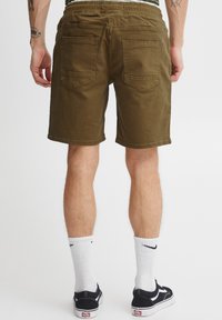 Pantaloni scurți verde măsliniu cu finisaj texturat, talie elastică și două buzunare la spate. Purtate cu adidași negri și șosete albe.