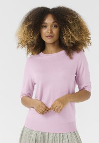 Gaišs rozā adīts īsām piedurknēm džemperis ar apaļu kakla izgriezumu un ribinātu apakšmalu, kombinēts ar plisētu gaišas krāsas svārku.