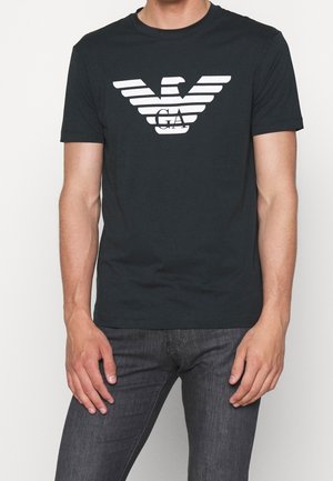 Homme portant un t-shirt noir à manches courtes avec un logo d'aigle stylisé blanc et le lettrage "GA", associé à un jean gris foncé.