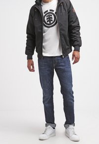 Czarna pikowana kurtka z kapturem, szara grafitowa koszulka, niebieskie dżinsy i białe trampki. Posiada kieszenie na zamek i ściągacze na rękawach.