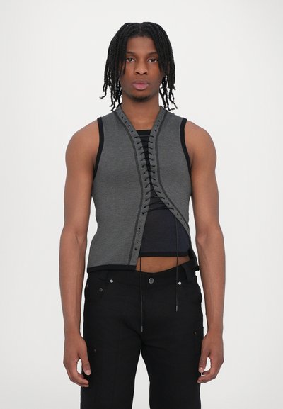 Gilet grigio senza maniche con design anteriore a laccio, caratterizzato da bordi neri a contrasto. Indossato sopra un top nero aderente con una silhouette corta.