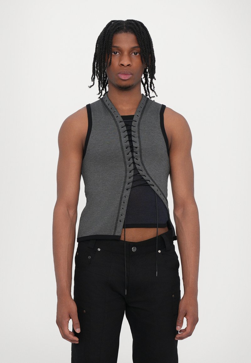 Gilet sans manches gris avec un design à lacets à l'avant, mettant en valeur des bords noirs contrastants. Porté par-dessus un haut noir ajusté avec une silhouette courte.