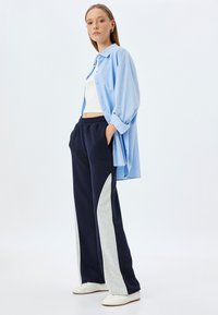 Lichtblauwe oversized button-up shirt, witte crop top, marineblauwe joggers met grijze zijkanten en witte sneakers. Gladde stof en relaxte pasvorm.