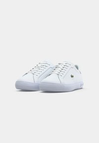 Des chaussures de sport blanches avec un dessus en cuir lisse, un embout rond, des lacets blancs et une semelle en caoutchouc translucide. Présente un petit logo vert.