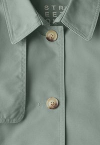 Jasnozielona kurtka z kołnierzykiem o gładkiej fakturze, z dwoma guzikami – jednym z zabawnym wzorem psa – na pionowej listwie.