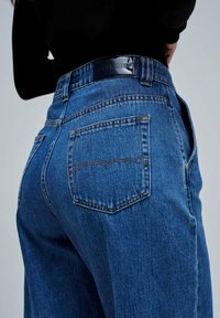 Jeans de mezclilla en un lavado azul medio con una cintura alta, con bolsillos traseros y detalles de costura. El logo de la marca está en la cinturilla.