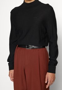 Schwarzer Pullover mit hochgeschlossenem Kragen, gerippten Bündchen und einem dezenten gestickten Logo. Kombiniert mit lockeren rostfarbenen Hosen und einem schwarzen Gürtel.