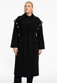 Zwarte wollen jas met een getailleerde taille en een strikceintuur. Voorzien van grote knopen op de schouders en twee voorkeurszakken. Lange mouwen en een reverskraag.