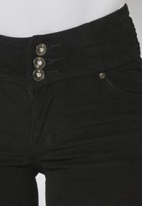 Pantalon en velours côtelé noir avec trois boutons métalliques décoratifs et une poche avant visible, porté avec un haut blanc.