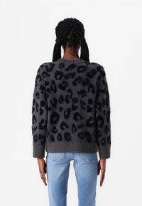 Maglione grigio oversize con stampa leopardata nera, texture a maglia e polsini a costine. Indossato con jeans azzurri chiari, che evidenziano un fit rilassato.