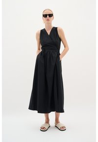 Schwarzes Wickelkleid mit V-Ausschnitt, ärmellosem Design und elastischem Taillenbund. Verfügt über zwei seitliche Taschen und fällt bis zur Mitte der Waden. Getragen mit leichten Sandalen.