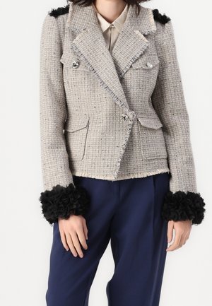 Blazer de tweed gris de doble botonadura con bordes deshilachados y puños negros con motivos florales, llevado sobre camisa beige y pantalones azul marino.