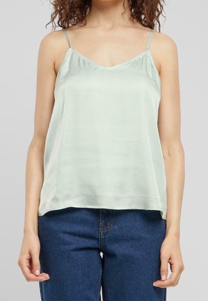 Kobieta ubrana w jasnozielony satynowy top na cienkich ramiączkach i ciemnoniebieskie dżinsy na neutralnym tle.