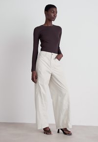 Hnedý top s dlhým rukávom v kombinácii so širokými krémovými nohavicami. Modelka má na sebe čierne sandále na podpätku, ktoré sa vyznačujú minimalistickým dizajnom. Hladké textúry.