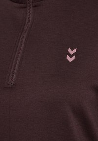 Burgundy zip-up topp i mjukt tyg med hög krage och en dekorativ rosa chevron-logotyp på vänster bröstområde.