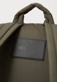 Intérieur d'un sac à dos en tissu vert olive avec un design matelassé, étiquette en cuir noir avec "VEE COLLECTIVE" en relief, texture lisse.