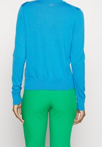 Blå stickad tröja med långa ärmar, ribbad mudd och nederkant. Har en liten logotyp på baksidan. Kombinerad med gröna tajta byxor.