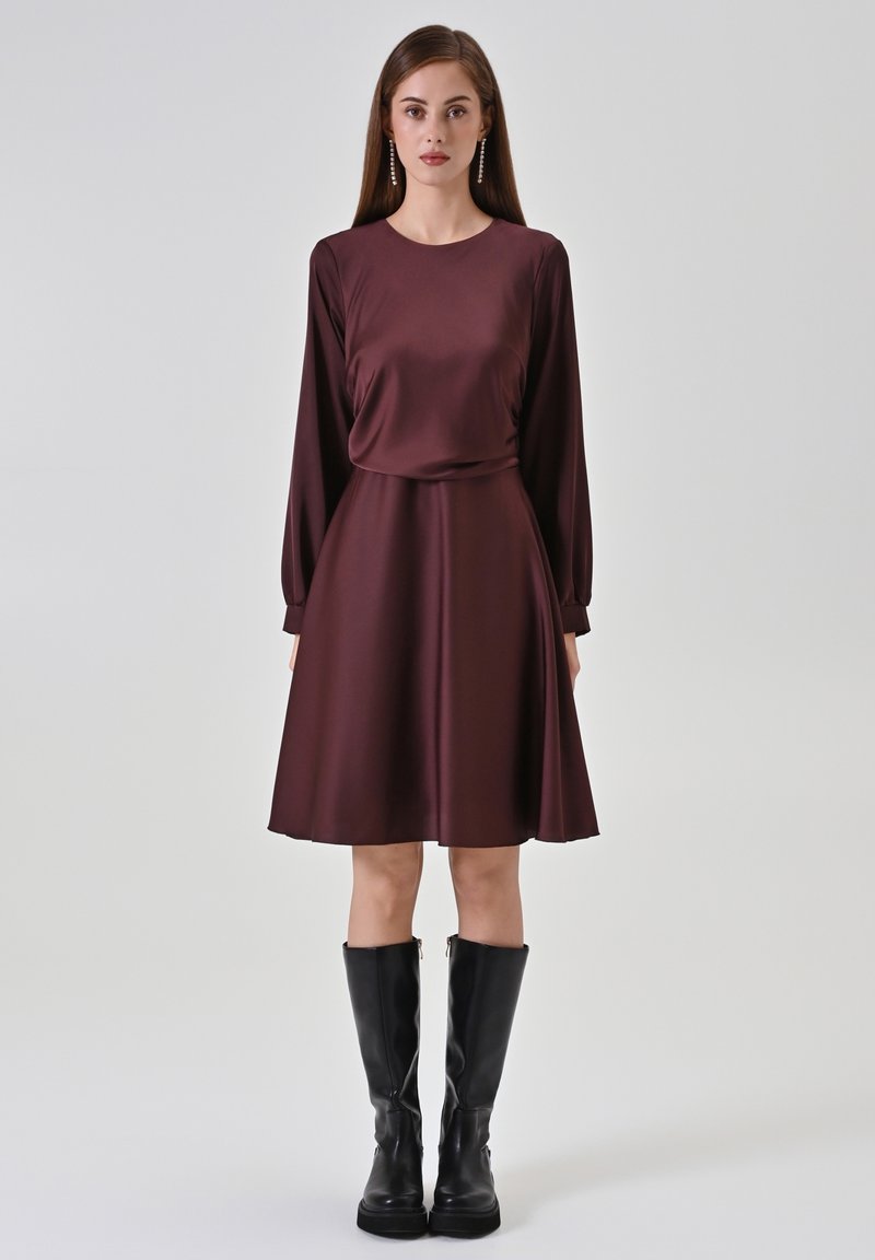 Bordeauxrotes Langarmkleid aus glattem Stoff mit taillierter Passform und ausgestelltem Rock, kombiniert mit kniehohen schwarzen Stiefeln.